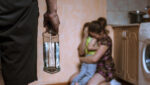 man holding alcohol bottle with woman and child hiding in corner of room